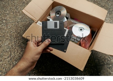 Two vintage floppy disks held in hand in front of a cardboard box full of old cd's and storage devices