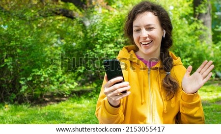 Smiling young woman making video call with mobile phone in green garden outdoors. Best friends having virtual conference, party online meeting use digital application for communication.Gesturing hi.