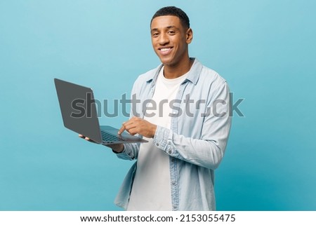 Optimistic african-american male student in casual jeans shirt using laptop pc isolated on blue background, smiling multiracial freelancer man looking enjoying remote work, advertising concept
