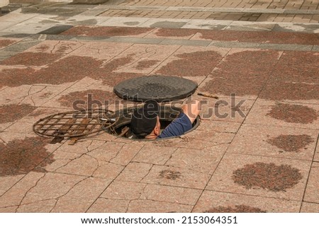 A worker climbs out of an open manhole in the road. Dangerous open unprotected manhole on the road. Accident with a sewer manhole in the city. The concept of repairing underground utilities. Royalty-Free Stock Photo #2153064351