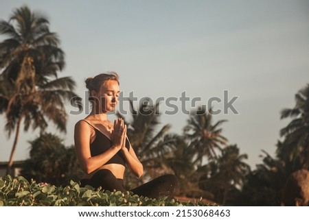 Young woman does yoga for healthy lifestyle on sea beach at palm trees background. Female performing sports exercises to restore strength and spirit. Yoga position on tropical climate Royalty-Free Stock Photo #2153068463
