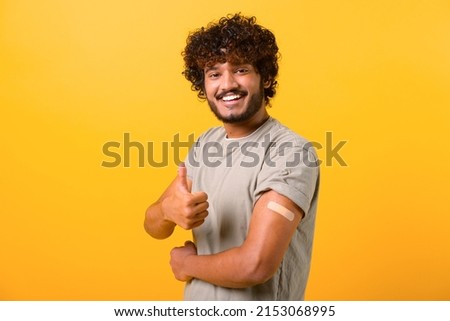 Smiling young curly Indian guy showing arm with band-aid after vaccine injection isolated on yellow background, vaccinated latino man showing thumb up