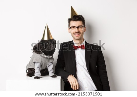 Image of handsome young man celebrating birthday with cute black pug in party costume and cone on head, standing over white background