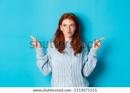 Winter holidays and people concept. Thoughtful redhead girl making decision, pointing fingers sideways, choosing between two ways, standing over blue background