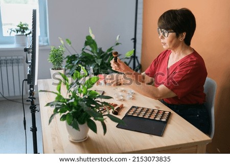 Middle-aged female beauty blogger or influencer with makeup product recording video in room at home. Using ring lamp and smartphone