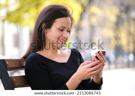 Young woman sitting on park bench browsing her cellphone outdoors on warm summer day. Communication and mobile connection concept