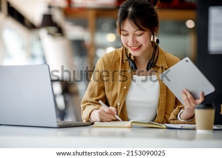 Young adult happy smiling Hispanic Asian student wearing headphones talking on online chat meeting using laptop in university campus or at virtual office. College female student learning remotely. Royalty-Free Stock Photo #2153090025
