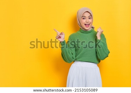 Excited beautiful Asian woman in green sweater pointing fingers aside at copy space isolated over yellow background