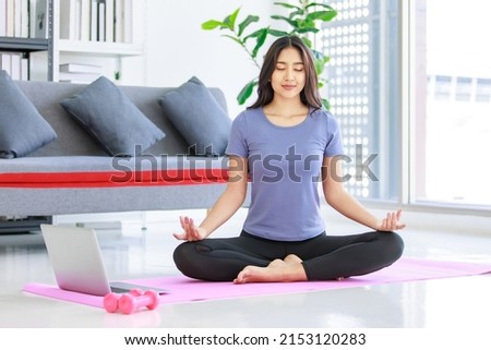 Asian young happy peaceful calm female model in casual sporty outfit sitting crossed legs in lotus position on yoga mat learning studying online meditation class via laptop computer in living room. Royalty-Free Stock Photo #2153120283