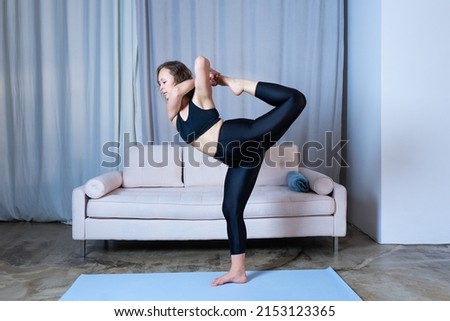 Young healthy beautiful woman in sports top and leggings practicing yoga at home, standing in pose on yoga mat