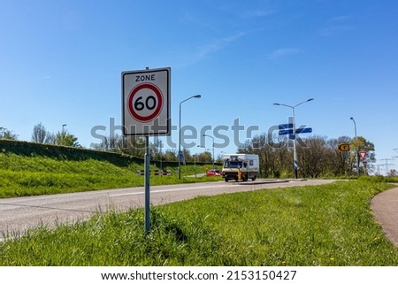 Rural road with a traffic sign: maximum speed zone 60, cars circulating, post with plates indicating different directions and trees in the background, sunny day in Netherlands