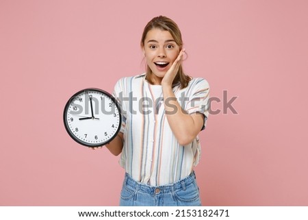Excited young blonde woman girl in casual striped shirt posing isolated on pastel pink background studio portrait. People emotions lifestyle concept. Mock up copy space. Hold clock put hand on cheek Royalty-Free Stock Photo #2153182471