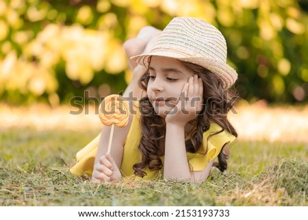 A young girl in a yellow suit and a hat with a lollipop in her hands outdoors in the park. High quality photo