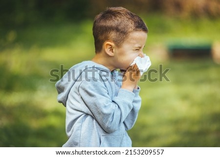 Child with pollen allergy. Boy sneezing and blowing nose because of seasonal allergy while sitting in a grass. Spring allergy concept. Flowering bushes and trees in background. Child allergy