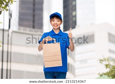 service and job concept - happy smiling delivery woman in blue uniform with takeaway food in paper bag showing thumbs up over city street background