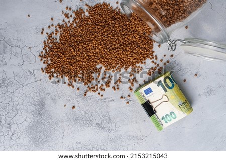 Buckwheat poured from glass jar and stack of money on gray background. Inflation,food crisis concept