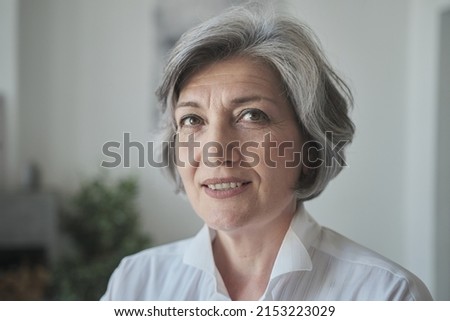 A confident, educated older lady executive manager stands in a bright contemporary workplace and appearing serious. A senior female professor or instructor headshot in university