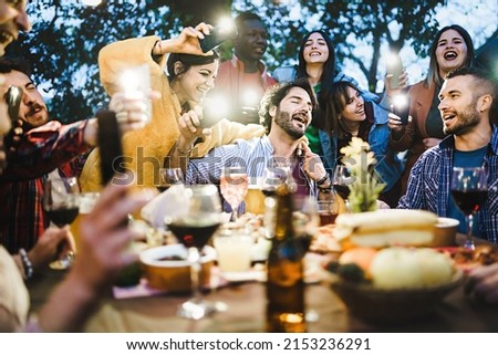 Group of drunk young people singing loud around the barbeque food table drinking wine and beers and making lights with smart phones torches - Friends get together at balcony rooftop party having fun