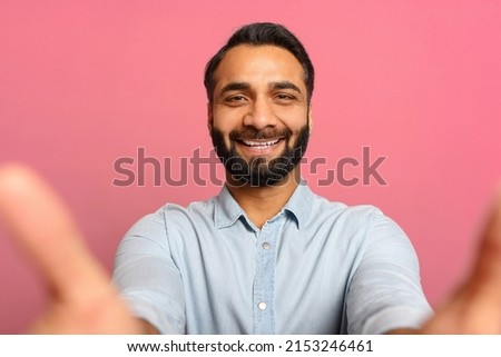 Portrait of bearded Indian handsome man taking selfie, looking at camera POV, brown-haired guy recording himself. Indoor studio shot isolated on pink background
