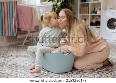 Loving mom takes care of little adorable daughter, little girl helps woman sort clothes for laundry, baby sits in bowl gives kiss to beloved mother.