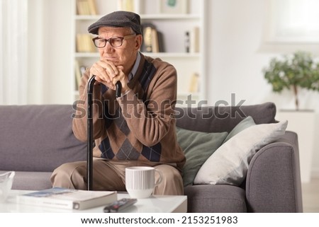 Grumpy elderly man sitting on a sofa with a walking cane at home Royalty-Free Stock Photo #2153251983