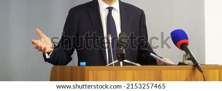 Man - politician, lawyer or official gesturing with his hands, holding documents and speaking from the podium in front of microphones. Speaker, orator, presenter or interviewee. No face. Web banner Royalty-Free Stock Photo #2153257465