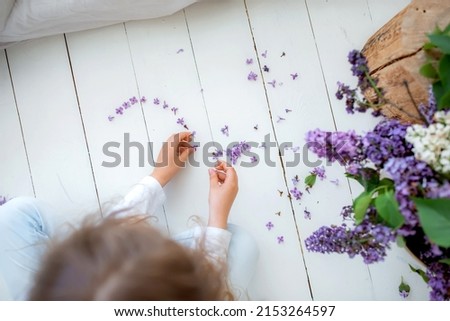 A little girl makes a figure of the heart of their lilac flowers on the white floor.