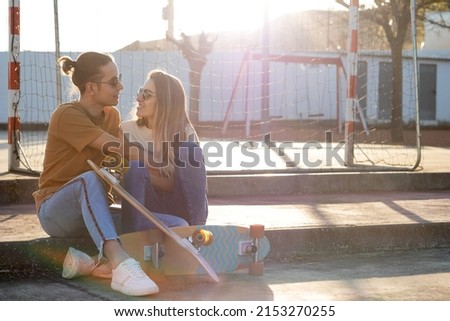In love couple looking to each other sitting in a park with two skate boards and sunset sun behind them