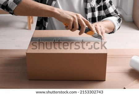 Man using utility knife to open parcel at wooden table, closeup