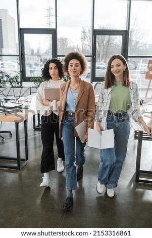 Happy interracial businesswomen with papers and laptop walking in office