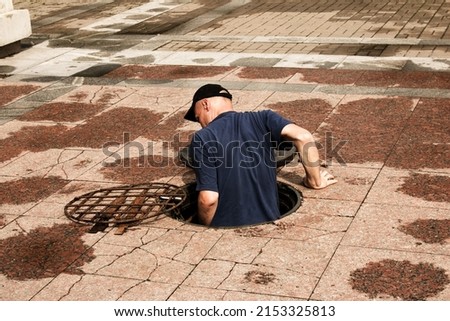 A worker climbs out of an open manhole in the road. Dangerous open unprotected manhole on the road. Accident with a sewer manhole in the city. The concept of repairing underground utilities. Royalty-Free Stock Photo #2153325813