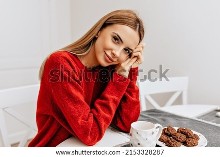 Lovable cute lady with light long hair wearing red seater is touching her chin and smiling while sitting in the kitchen at home with tea. High quality photo
