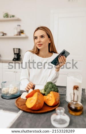 Appealing pretty lady with long hair dressed white shirt is sitting in the kitchen and searching recipe of healthy dishes. Woman preparing dinner from fresh vegetables. High quality photo