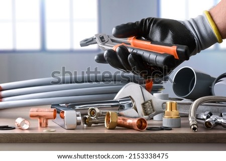 Sample of plumbing materials and tools on workbench and hand showing parrot beak pliers. Top view. Horizontal composition. Royalty-Free Stock Photo #2153338475