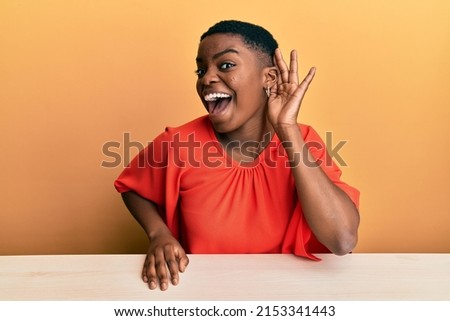 Young african american woman wearing casual clothes sitting on the table smiling with hand over ear listening and hearing to rumor or gossip. deafness concept.  Royalty-Free Stock Photo #2153341443