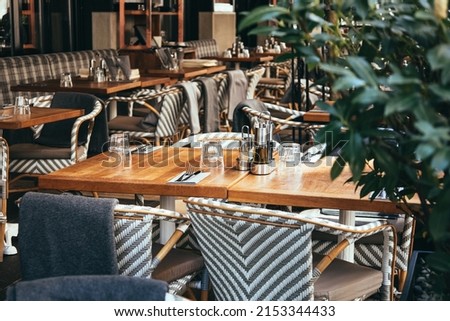 Outdoor empty coffee and restaurant terrace with wooden tables, vintage chairs and warm plaids. Green cafe terrace on the pedestrian street in Europe. A set table is waiting for customers. Royalty-Free Stock Photo #2153344433