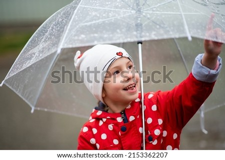 A cute little girl in a red cape, red boots and a white hat jump in puddles and has a fun.The girl has a transparent umbrella in her hands. Happy childhood. Early spring. Emotions.
