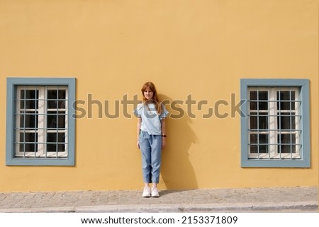 A red-haired teenage girl in blue clothes stands against a yellow wall with blue windows. Summer on a sunny day.