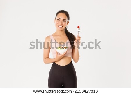 Sport, wellbeing and active lifestyle concept. Happy female athlete, asian fitness girl in activewear holding salad and tomato on fork, smiling, workout coach share diet and perfect body secrets