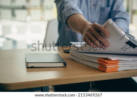 An employee sits at a table full of paper. stamp on the pile of unfinished documents A young account manager's secretary works in the office among piles of paperwork on the table. Royalty-Free Stock Photo #2153469267