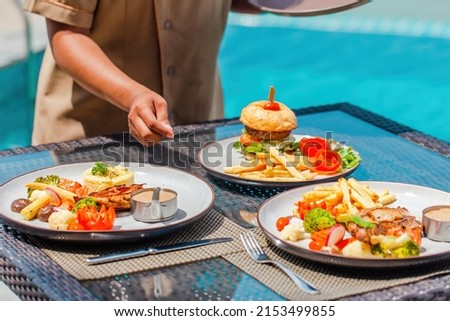 Waitress hand serving lunch in restaurant at luxury hotel. Asian woman waiter put delicious food plates on table in outdoors pool cafe in tropical resort. Summer travel, holidays, vacations concept. Royalty-Free Stock Photo #2153499855