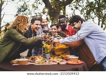Company of cheerful friends having fun toasting pic nic in farmhouse countryside - life style concept with multigenerational happy friends hands up toasting glasses of red wine in the back garden