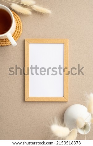 Top view vertical photo of wooden photo frame ceramic vase with white lagurus flowers and cup of tea on rattan serving mat on beige background with copyspace