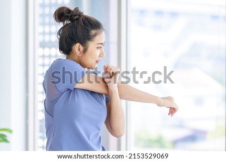 Asian young beautiful happy healthy female sport girl in casual sporty outfit standing smiling holding hand up stretching arm and body exercising training routine yoga pilates practice in living room.