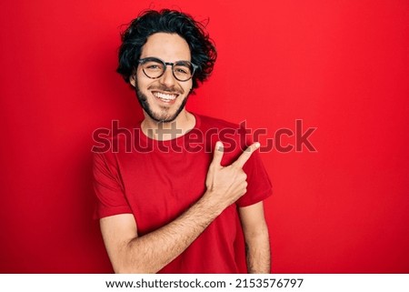 Handsome hispanic man wearing casual t shirt and glasses cheerful with a smile of face pointing with hand and finger up to the side with happy and natural expression on face 