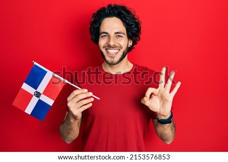 Handsome hispanic man holding dominican republic flag doing ok sign with fingers, smiling friendly gesturing excellent symbol 