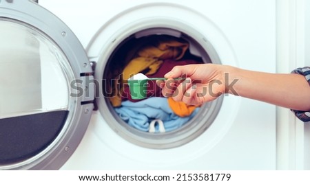 A woman's hand holds a cap with washing powder against the background of a washing machine with bright things put into it. The girl washes and wringes out things in the laundry room at home. Royalty-Free Stock Photo #2153581779
