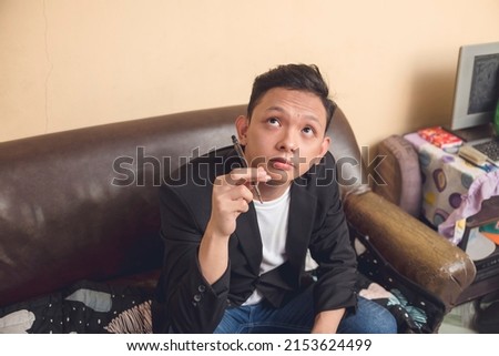 A young man sitting on a couch with a pen. Busy thinking what might he write on the essay. Constructing a sentence that is answering a question.