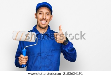 Bald man with beard holding roller painter smiling happy and positive, thumb up doing excellent and approval sign 