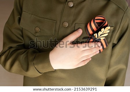St. George's ribbon is fixed on the chest of boy, his hand on the heart place in the military uniform. Concept of memory, symbol of Victory Day. The continuity of generations.
